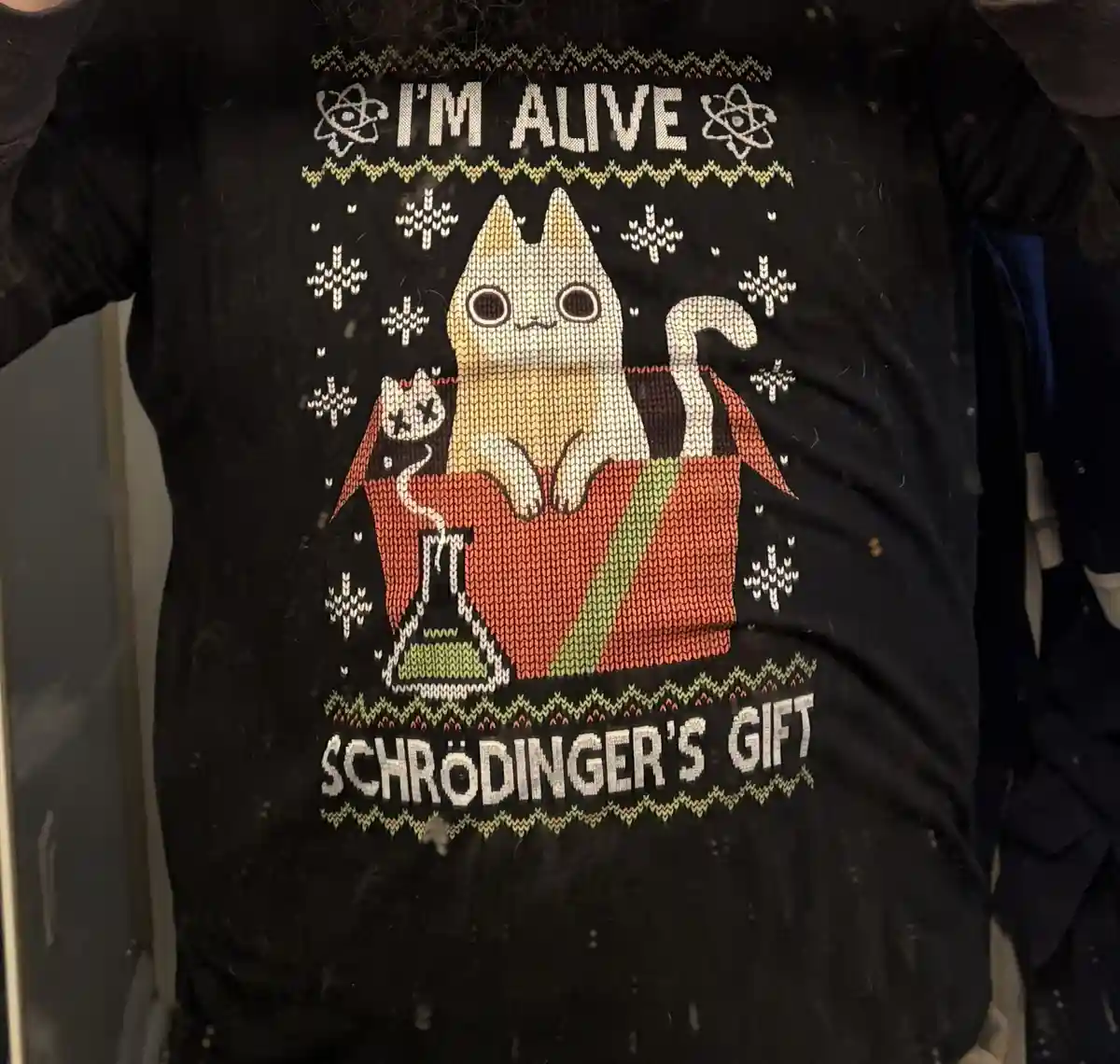 A black, knitted sweater features a cartoon cat sitting atop a red box with a green stripe, next to a beaker with smoke. The…