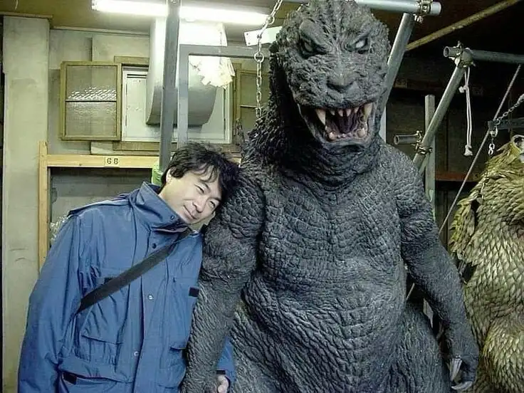 A man in a blue work jacket leans his head against a large, grey Godzilla costume, smiling tiredly. The costume is suspended…