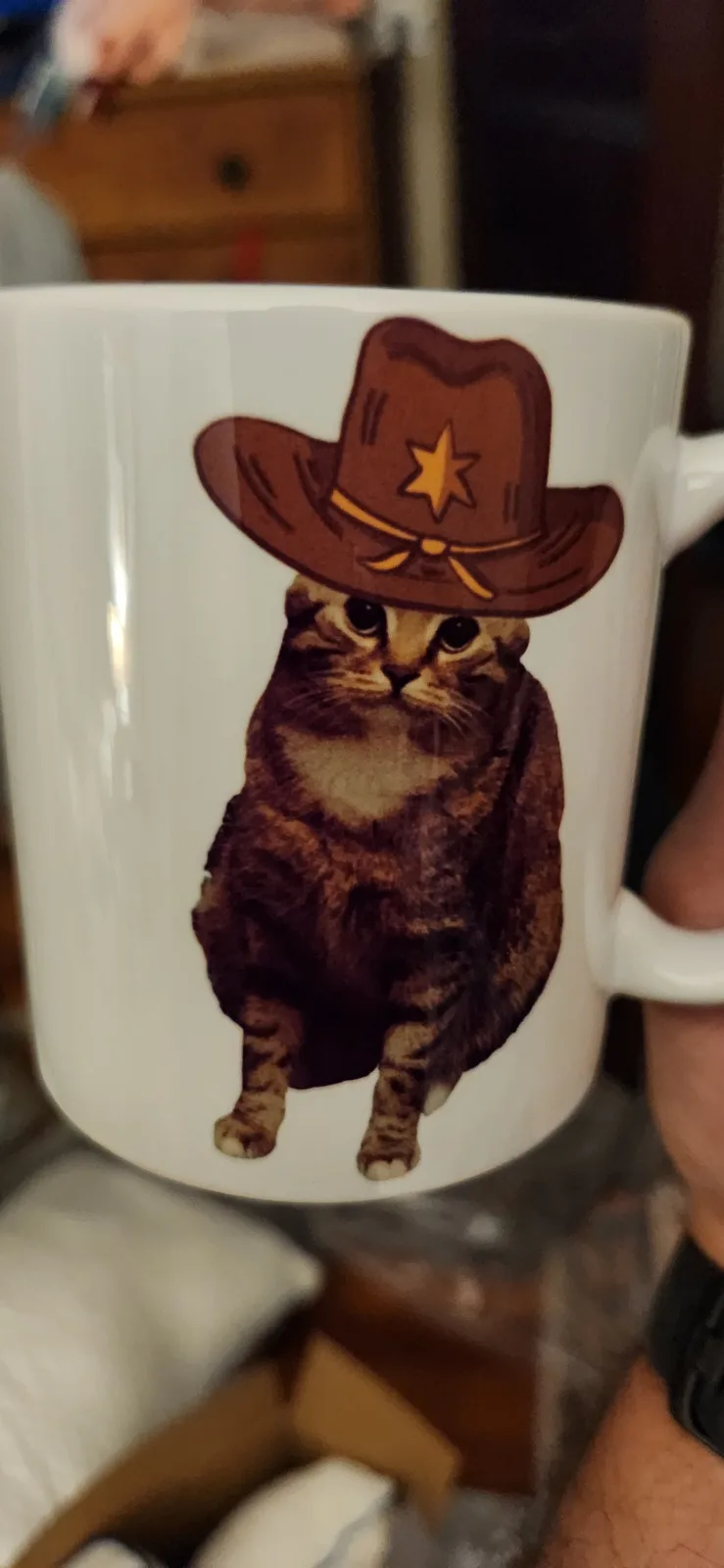 A white ceramic mug is held in someone's hand, featuring a printed image of a tabby cat wearing a brown cowboy sheriff's hat…