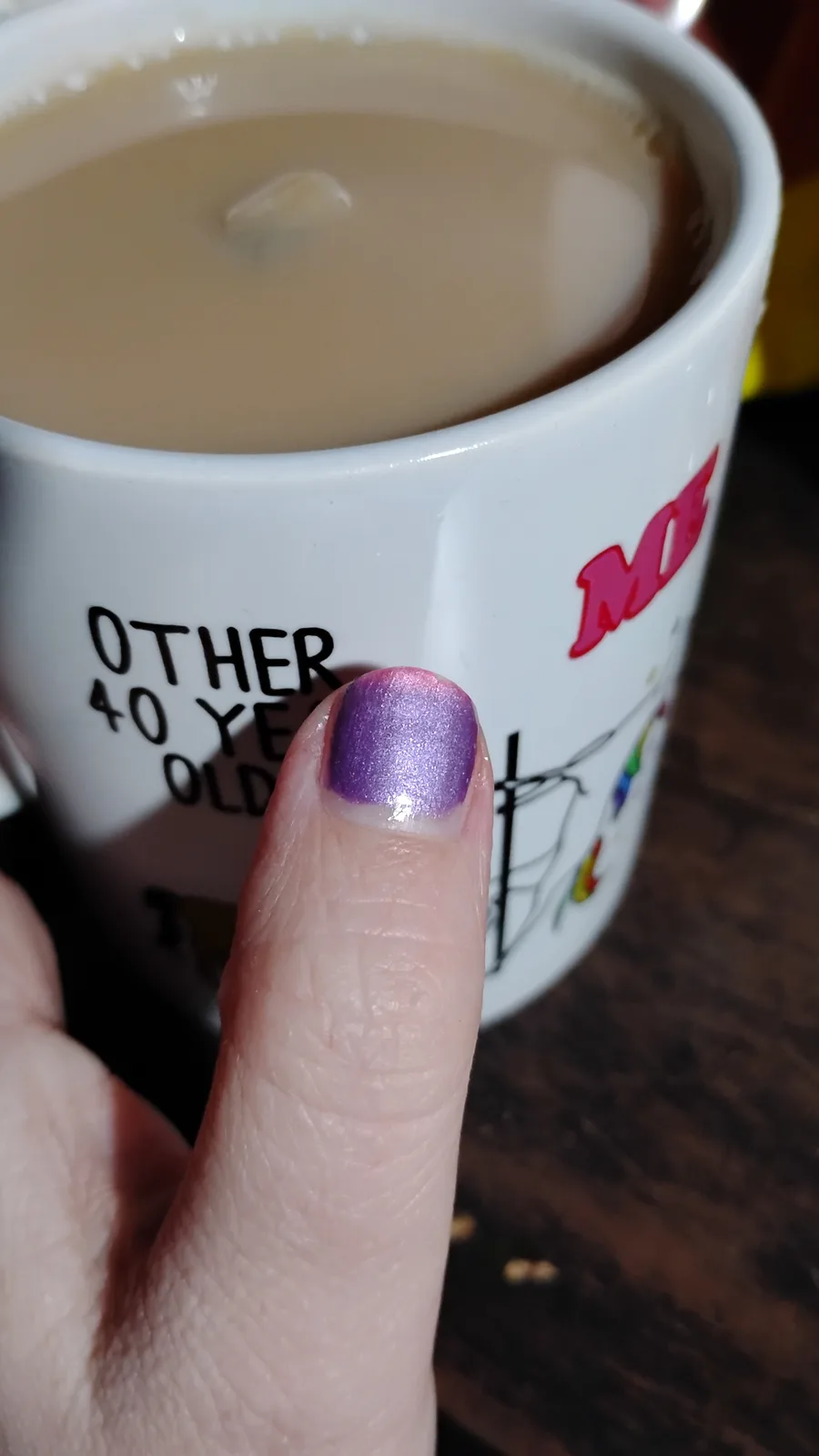 A close-up photo shows a person's thumb with sparkly purple nail polish, holding a white mug filled with light brown liquid…