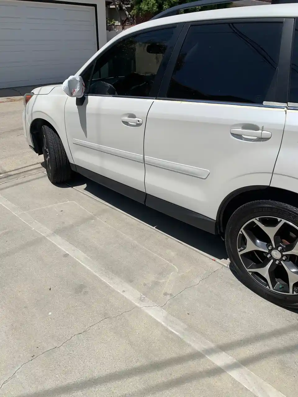 A white SUV is parked in a driveway with a white plastic bag covering its side-view mirror, implying the car was just…