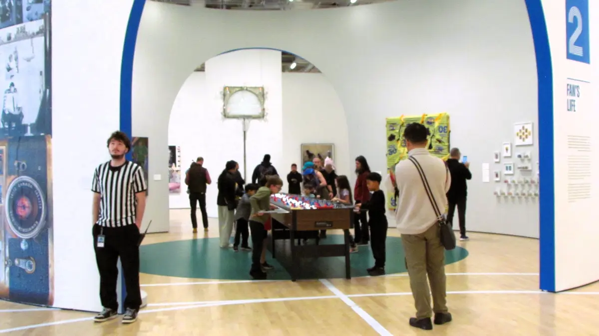 A man in a black-and-white striped referee shirt stands in a museum-like space with white walls and wooden floors marked…