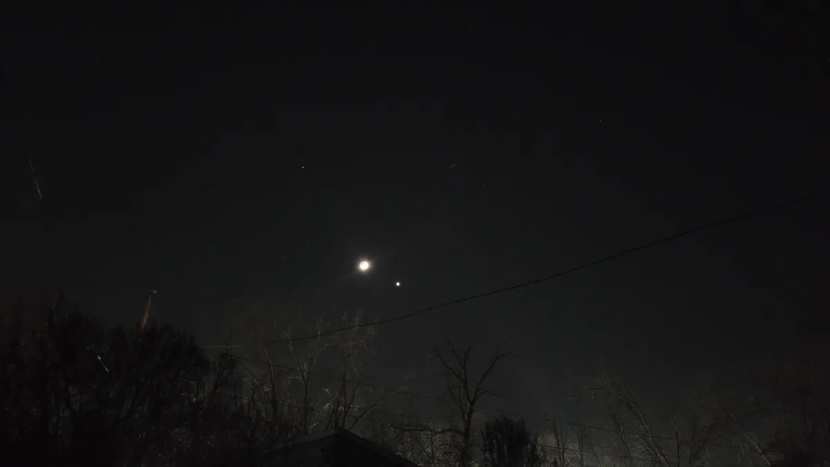 A night sky photo showing the moon and a smaller star or planet nearby, with silhouettes of bare trees and power lines…