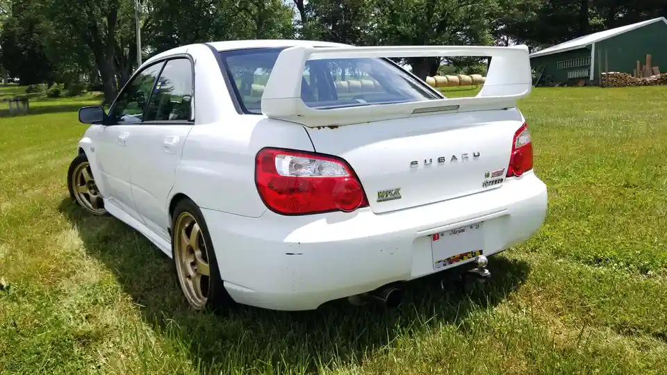 A white Subaru WRX STI sedan is parked on a grassy field with trees and a barn in the background. The car has a large rear…