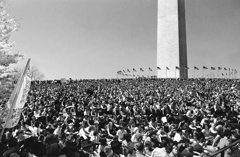 A black-and-white photo shows a massive crowd of people sitting on the grass in front of the Washington Monument, with…