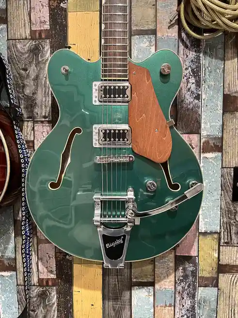 A green semi-hollow electric guitar with a Bigsby tailpiece is mounted on a wall of colorful, weathered wooden planks. The…