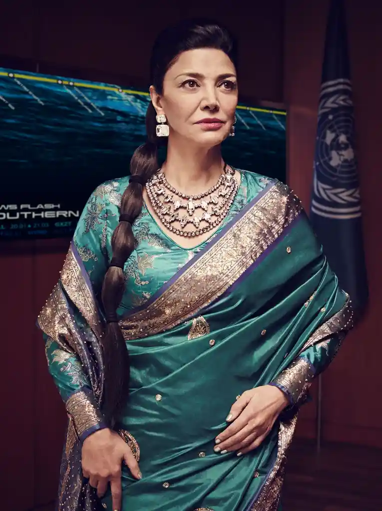 A woman in a green and gold sari with a long braid, adorned with heavy jewelry, stands in front of a screen displaying a…