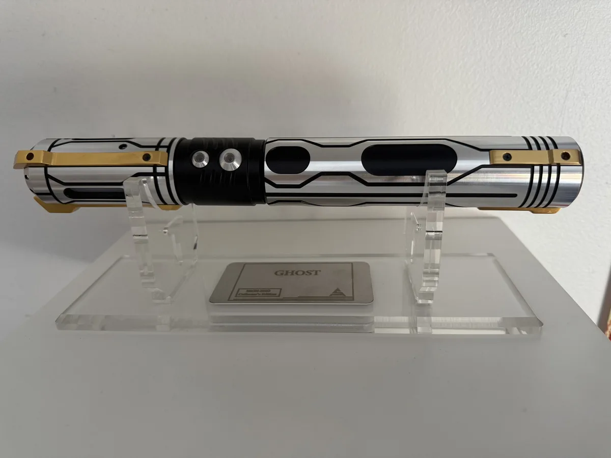 A silver and gold lightsaber hilt with black accents is displayed horizontally on a clear acrylic stand against a white…