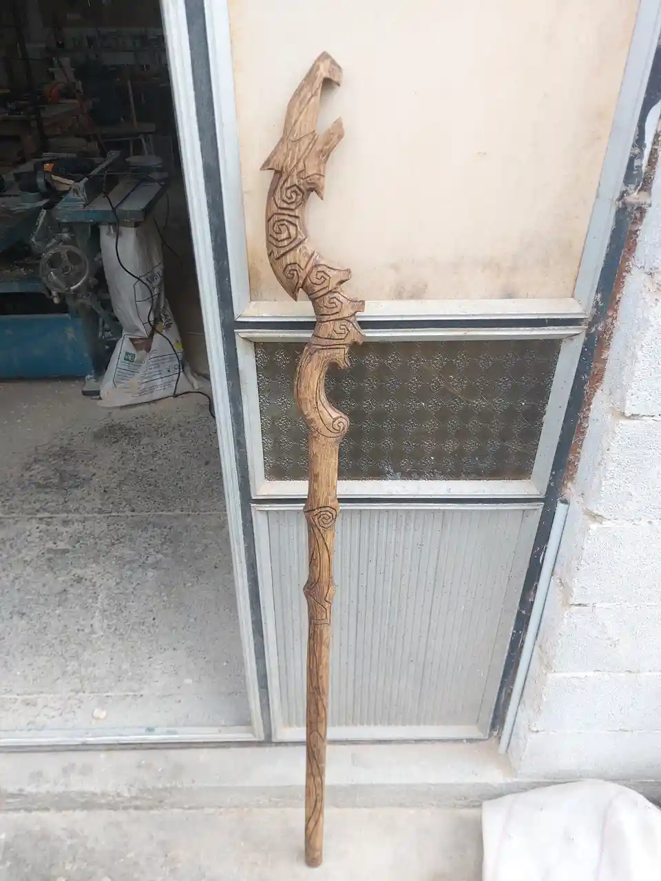 A highly detailed, hand-carved wooden staff with an ornate dragon head at the top leans against a glass-paneled door in what…