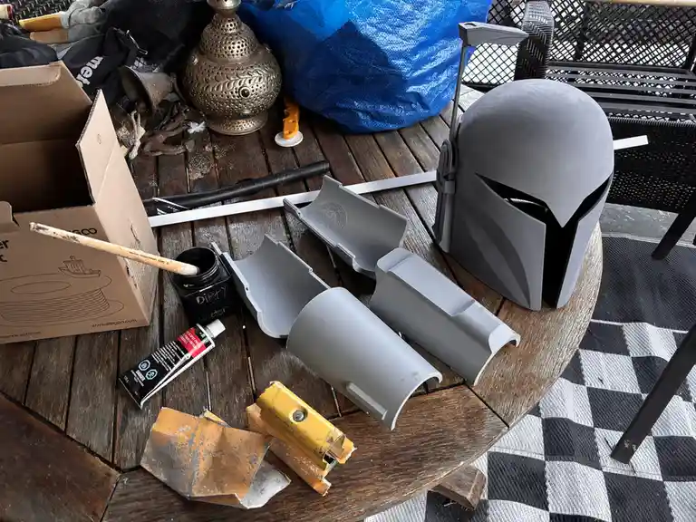 A wooden table is cluttered with crafting supplies including a partially assembled gray Mandalorian helmet, paint, glue, and…