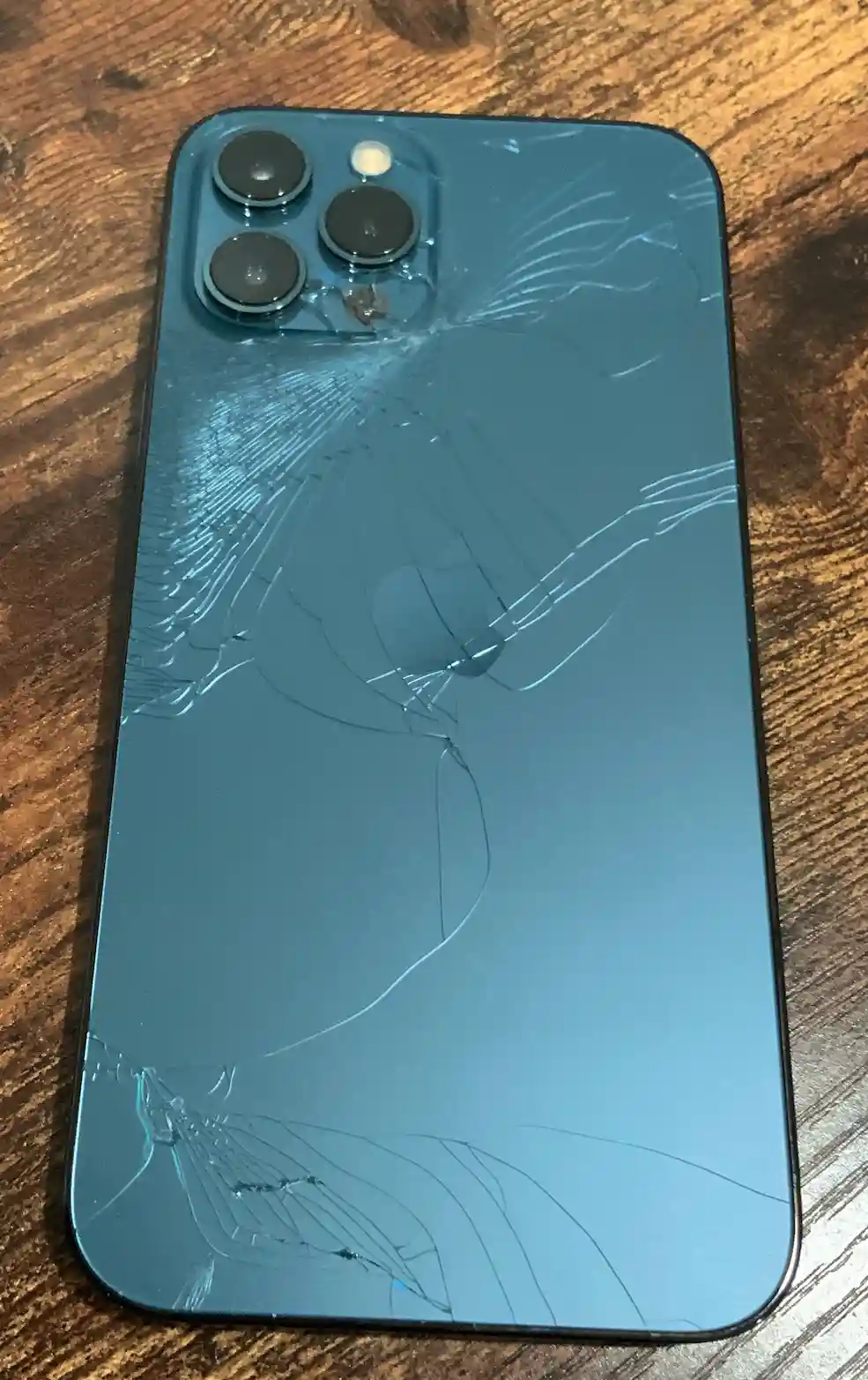 A blue smartphone with a shattered glass back lies on a wooden surface, cracks radiating from the camera module. The…