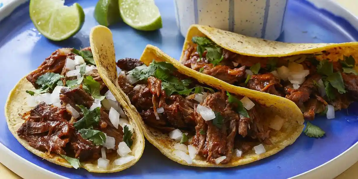 Three soft tacos filled with shredded meat, chopped onions, and cilantro sit on a blue plate, accompanied by lime wedges and…