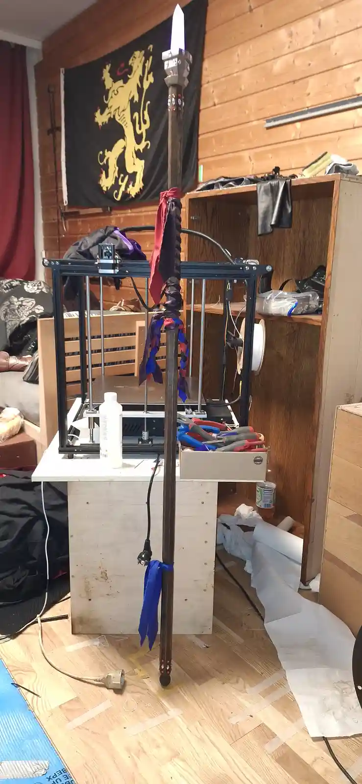 A 3d printer sits on a wooden stand in a cluttered room, with a large, ornate sword standing upright beside it. The sword…