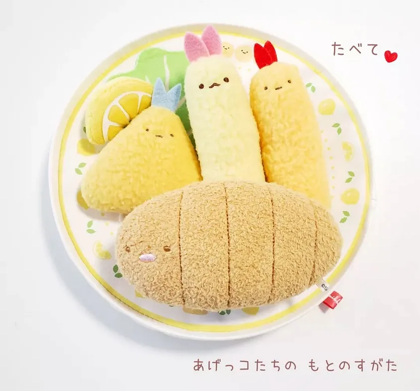 A white plate holds four cute, fluffy plush food items: a sliced lemon, a pear, a banana with pink bunny ears, and a bread…
