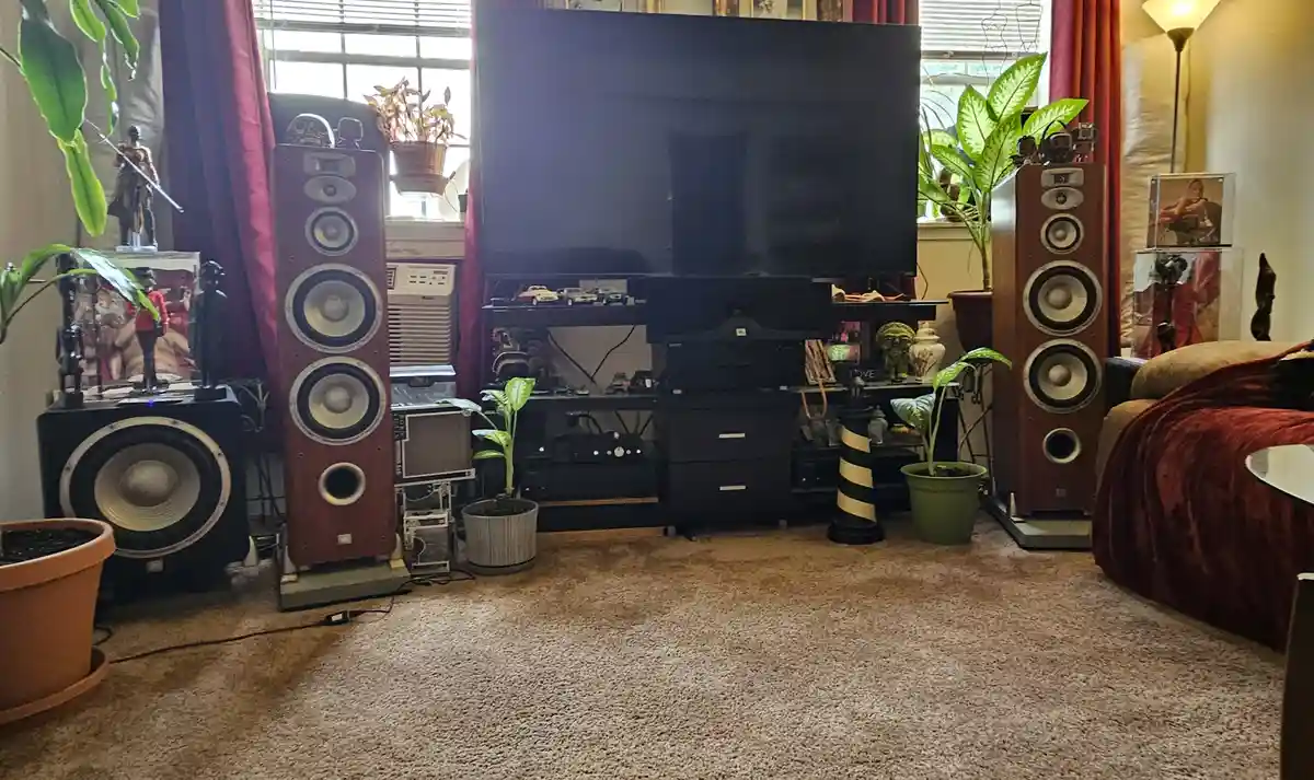 A living room features a large TV flanked by tall, expensive-looking floor-standing speakers and a subwoofer, surrounded by…