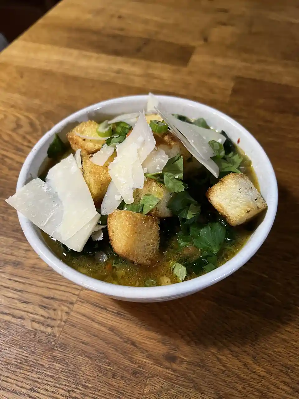 A white ceramic bowl filled with a greenish-yellow broth, topped with golden-brown croutons, fresh green herbs, and large…