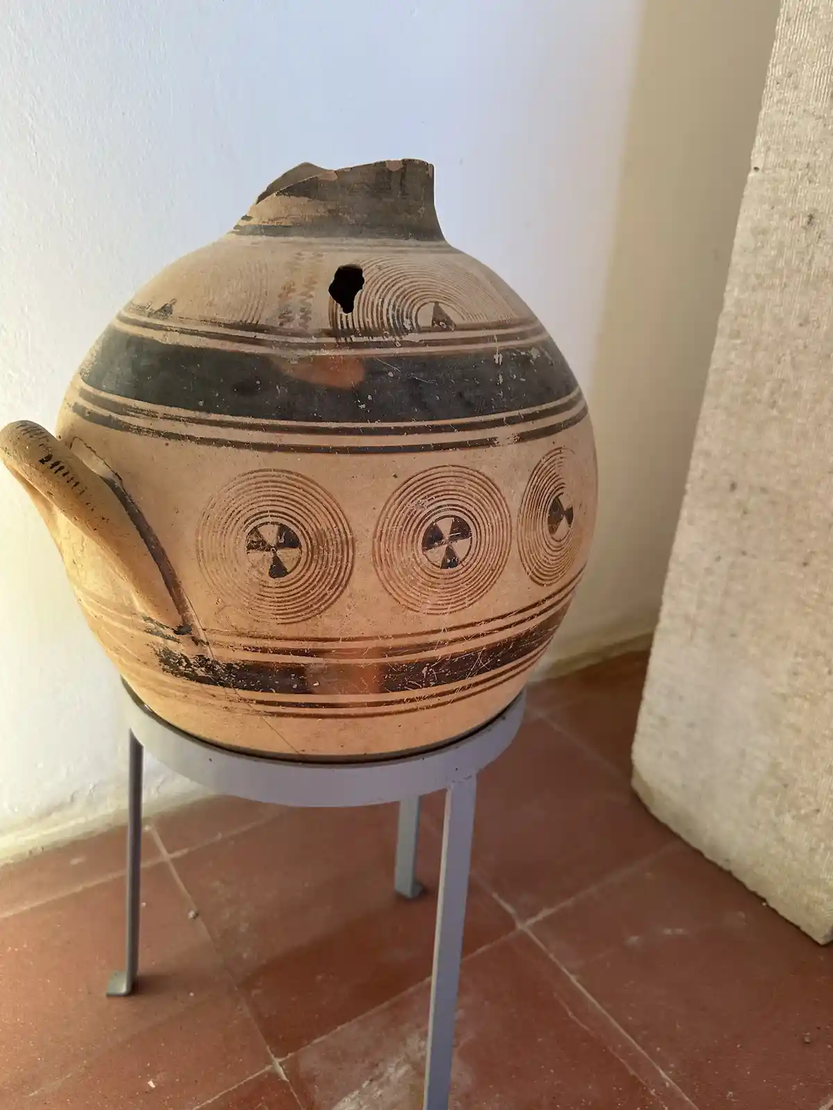 a weathered, terracotta-colored ancient pottery vessel with a handle and decorative concentric circle patterns sits on a…