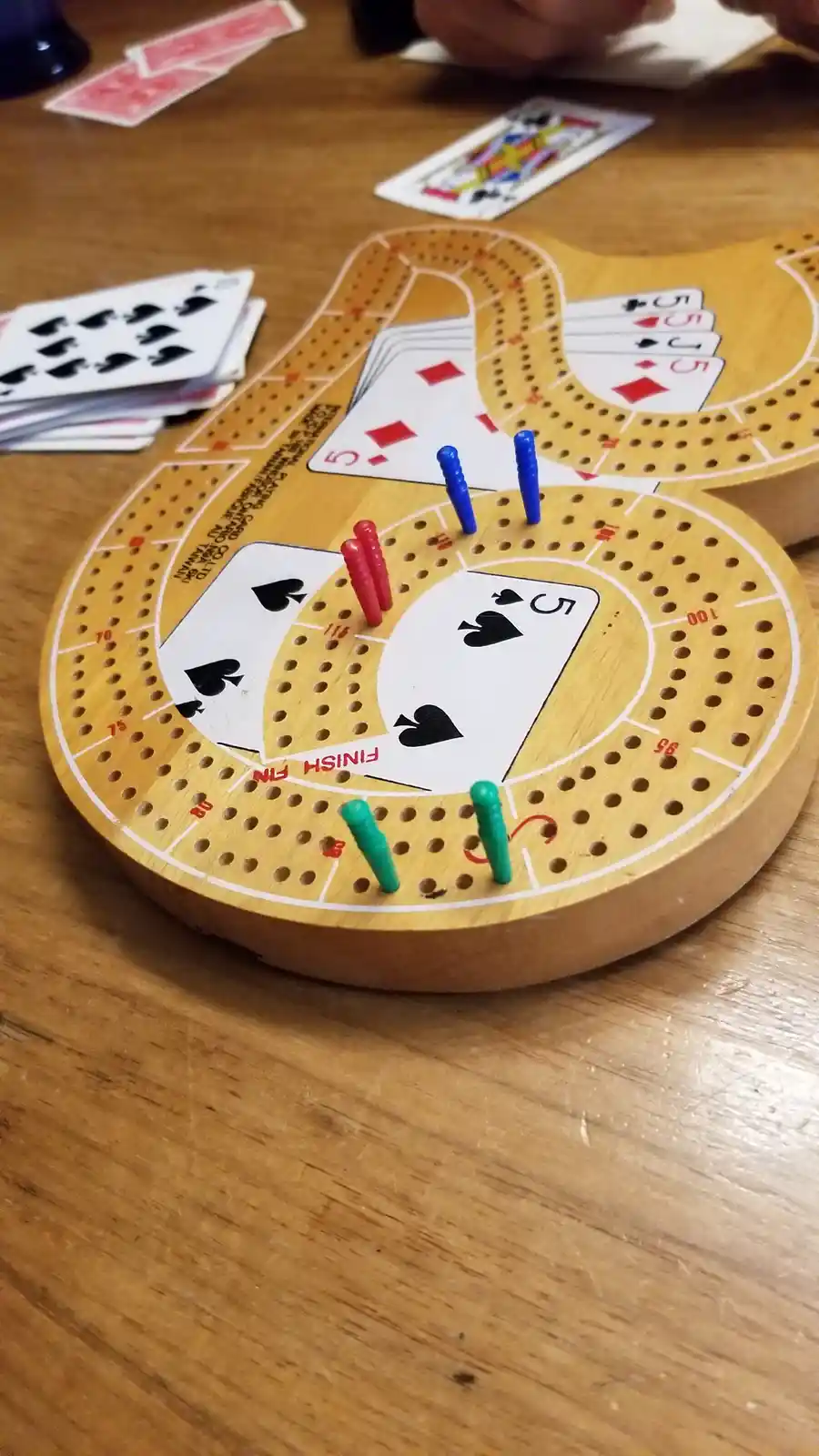 A close-up of a wooden cribbage board with playing cards placed on the scoring tracks and colored pegs inserted into holes…