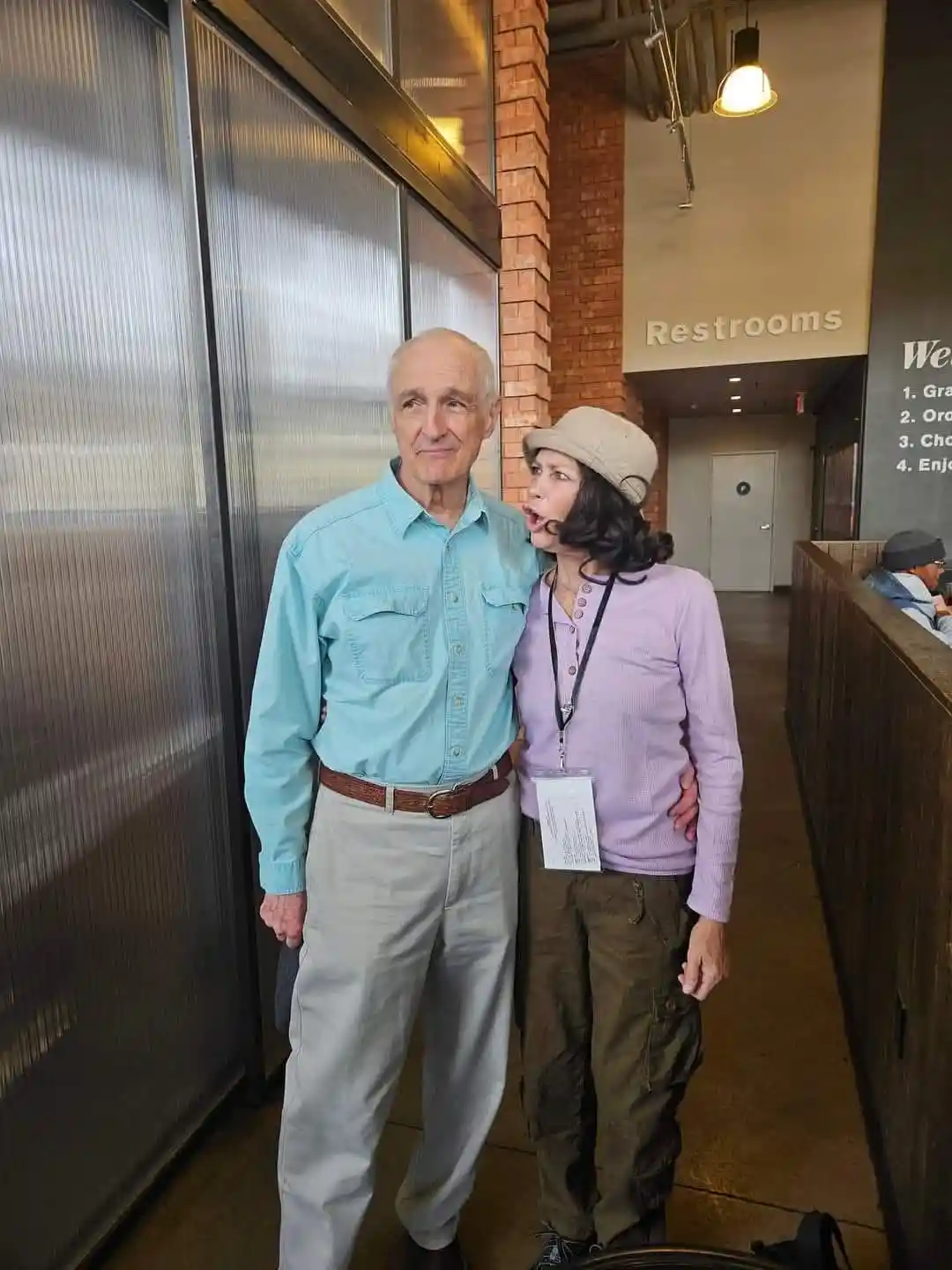 An older man and woman pose for a photo in a public building hallway. The woman, wearing a beige hat and purple shirt, leans…