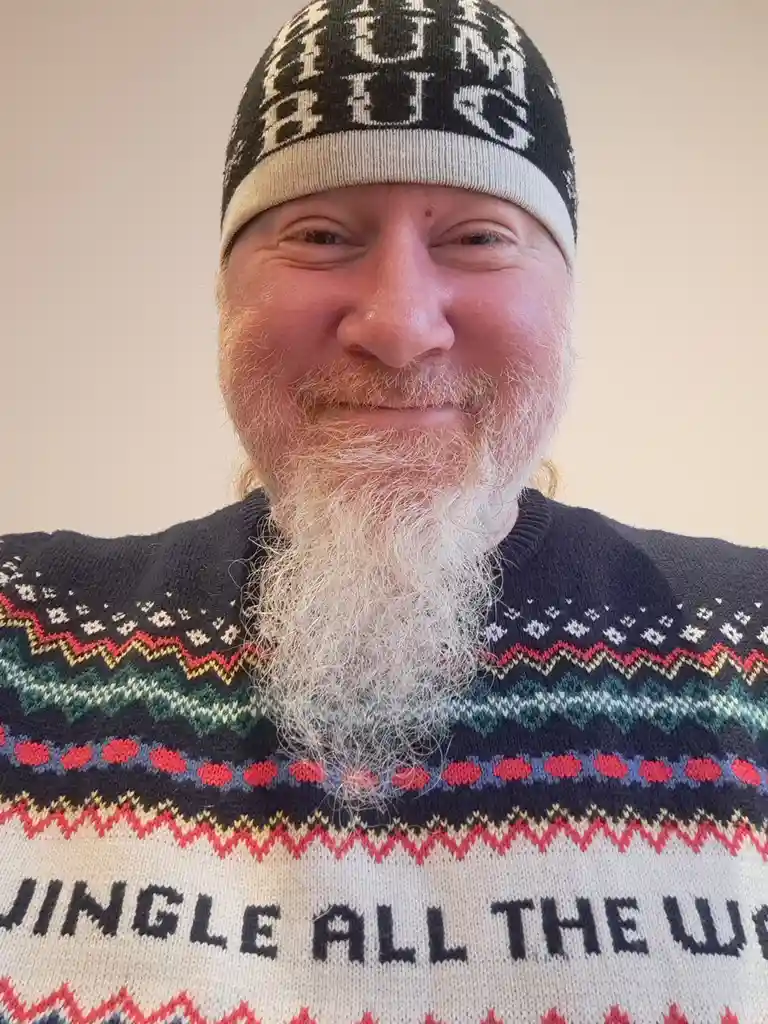 A man with a long white beard and a knit beanie smiles at the camera while wearing a colorful, patterned sweater that has…