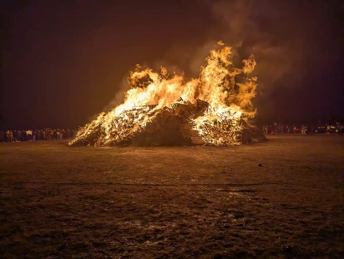 A large bonfire made of wooden debris burns intensely at night in an open field, with crowds of people standing at a…