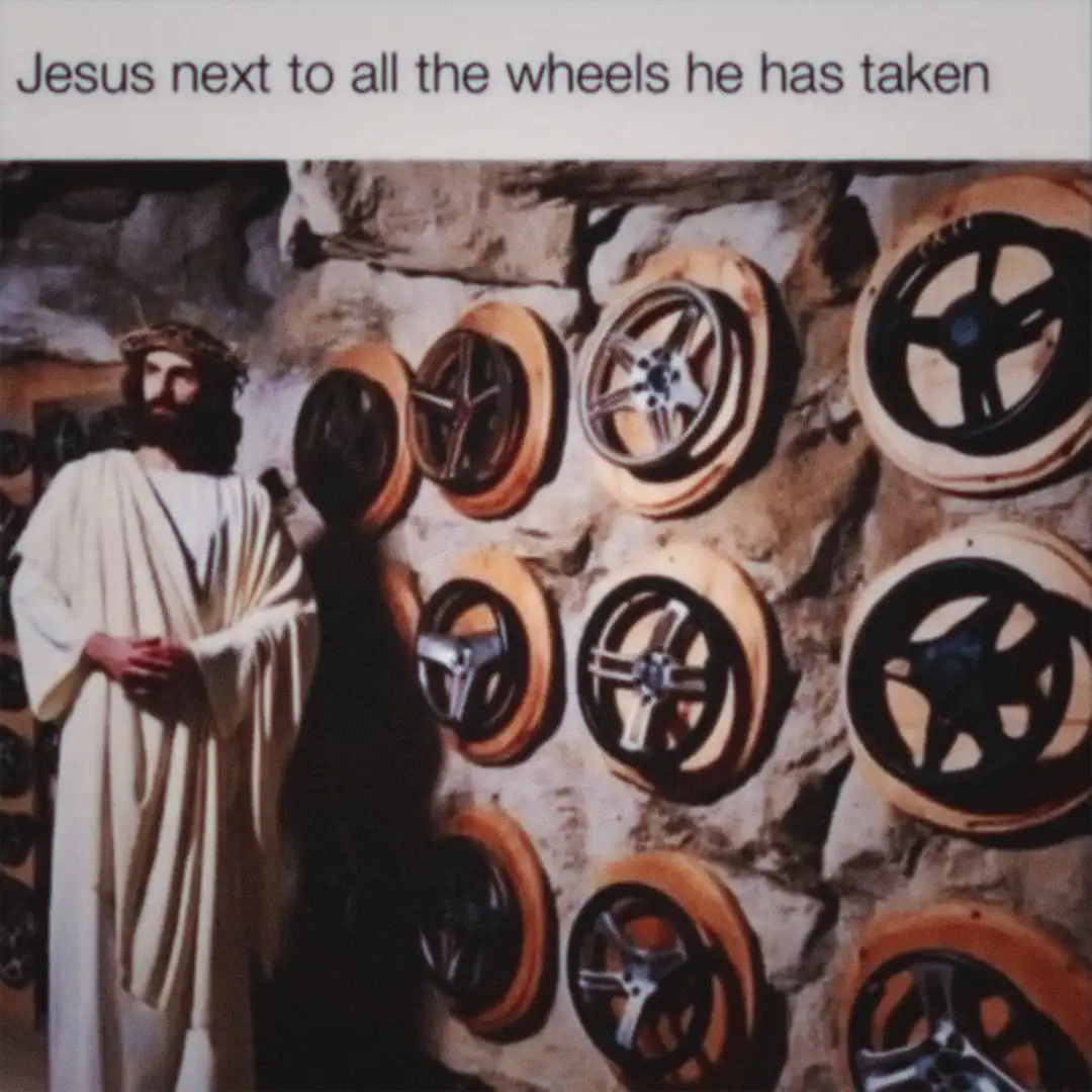A statue of Jesus Christ in a white robe and crown of thorns stands beside a stone wall covered with numerous car wheels…