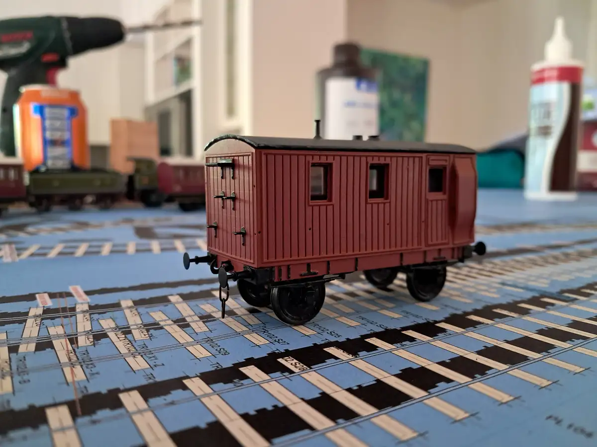 A red model train car sits on a printed track layout, surrounded by hobby tools like a drill and glue. The overlaid text…