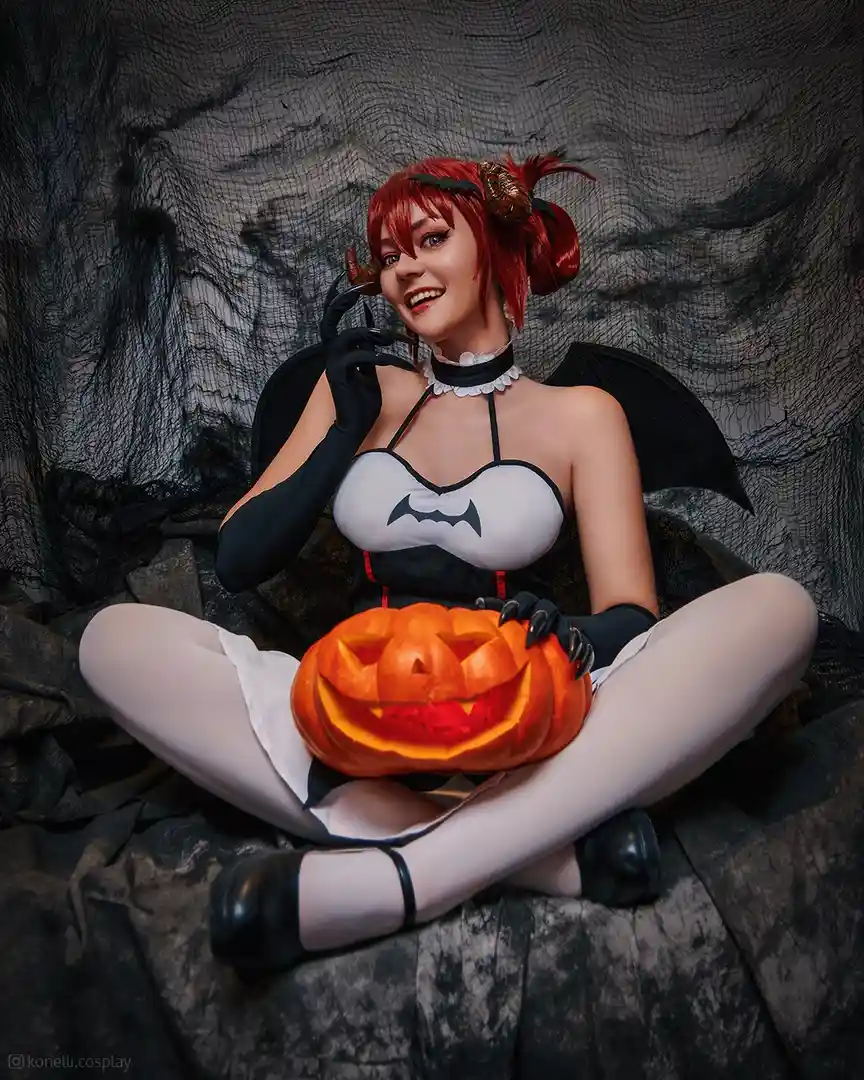 A person in a bat-themed Halloween costume with red hair, black wings, and a white top with a bat symbol, sits cross-legged…