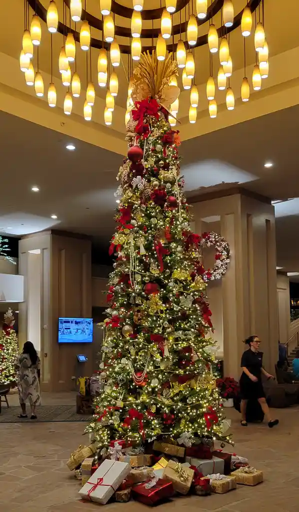 A large, elaborately decorated christmas tree stands in a hotel lobby, adorned with red and gold ornaments, lights, and a…