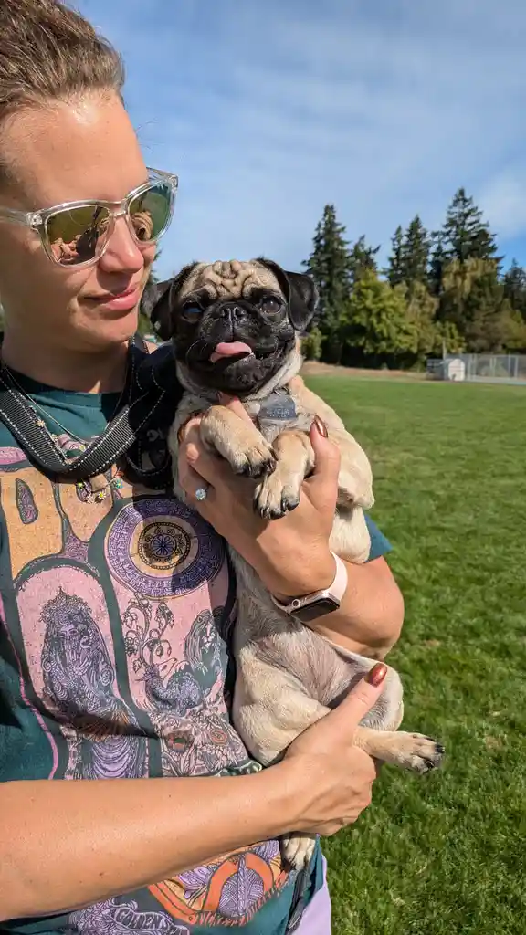 A person wearing sunglasses and a patterned shirt holds a pug puppy outdoors on grass. The pug has its tongue out and looks…