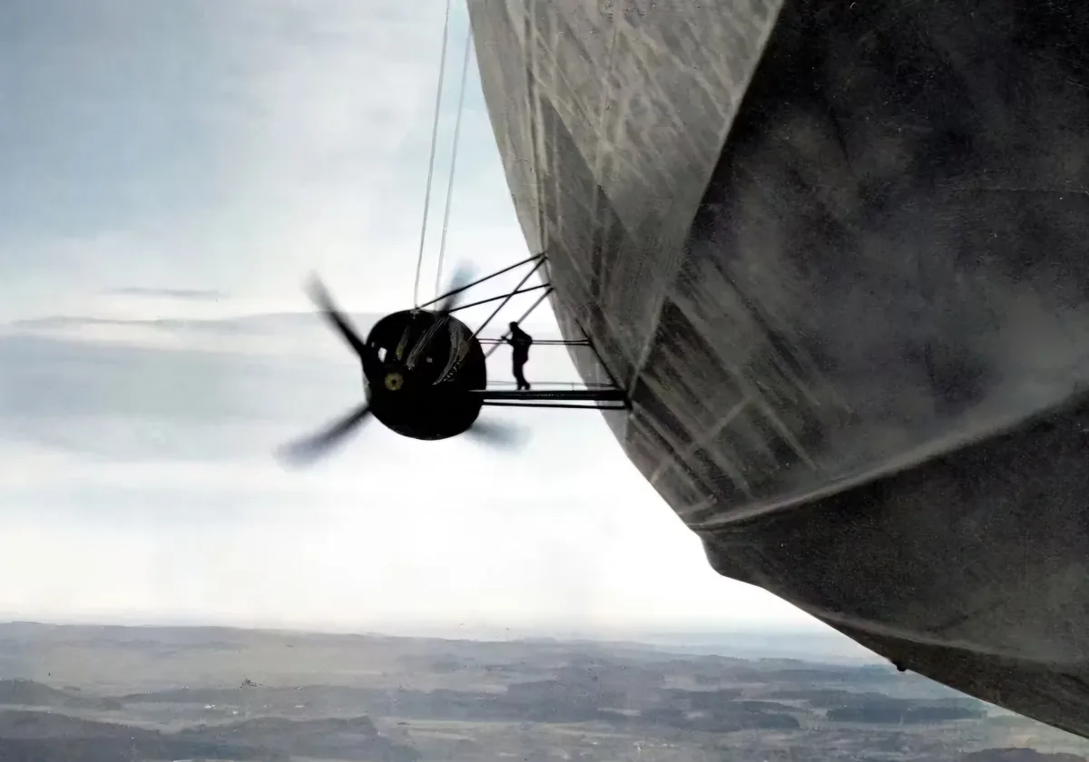 A person is silhouetted standing on a narrow metal platform attached to the side of a large, grey blimp, with a spinning…