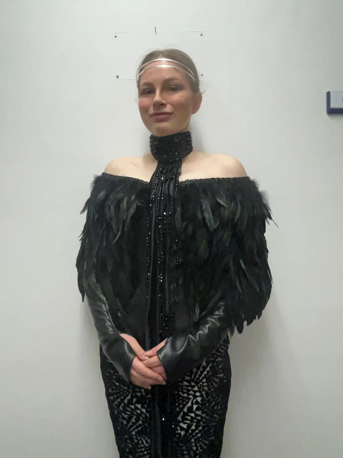 A woman stands against a plain white wall wearing an elaborate black outfit featuring a high-necked, feathered…