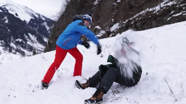 Two people in winter gear are engaged in a snowball fight in a snowy mountainous area. One person in a blue jacket and red…