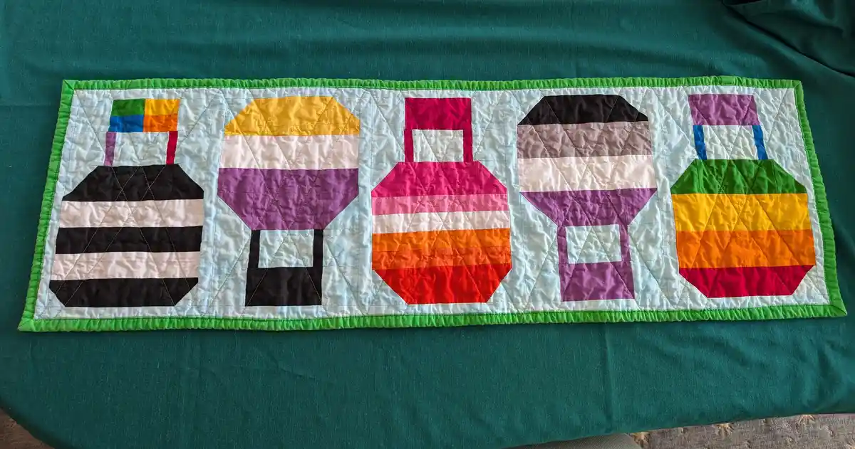 A handmade quilted runner displays five distinct pride flags, each rendered in colorful fabric blocks with visible…