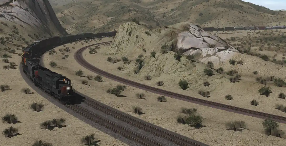 A long freight train curves through a dry, hilly desert landscape with sparse vegetation. The train is on a double-track…