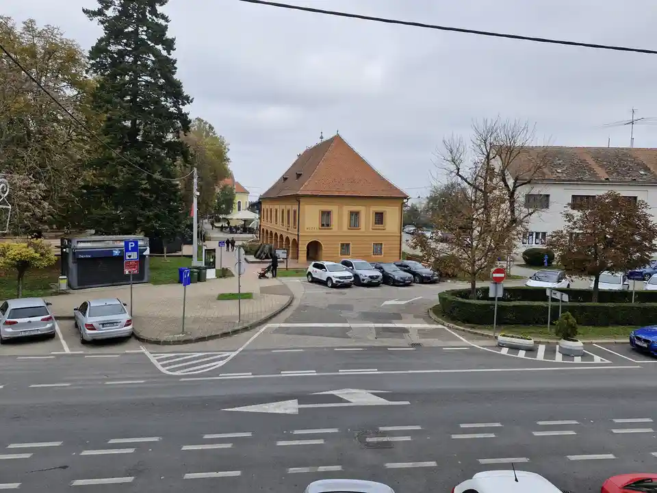 A street view of a yellow building with a red roof labeled 'MUZEJ' (museum) in Croatian, surrounded by parked cars and trees…