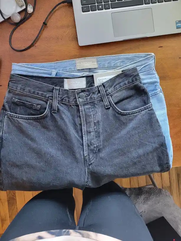 Two pairs of Everlane jeans are folded and stacked on a wooden desk, with a laptop and crystals nearby. The top pair is…
