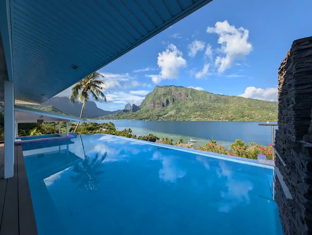 A serene, infinity-edge swimming pool with crystal blue water reflects a bright blue sky with fluffy white clouds. The pool…