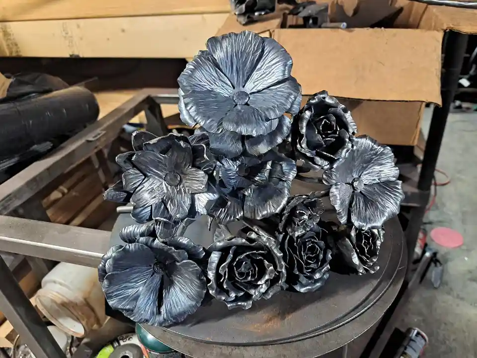 A metal sculpture of various flowers, including roses and daisies, crafted from dark, shiny metal, sits on a workbench in a…