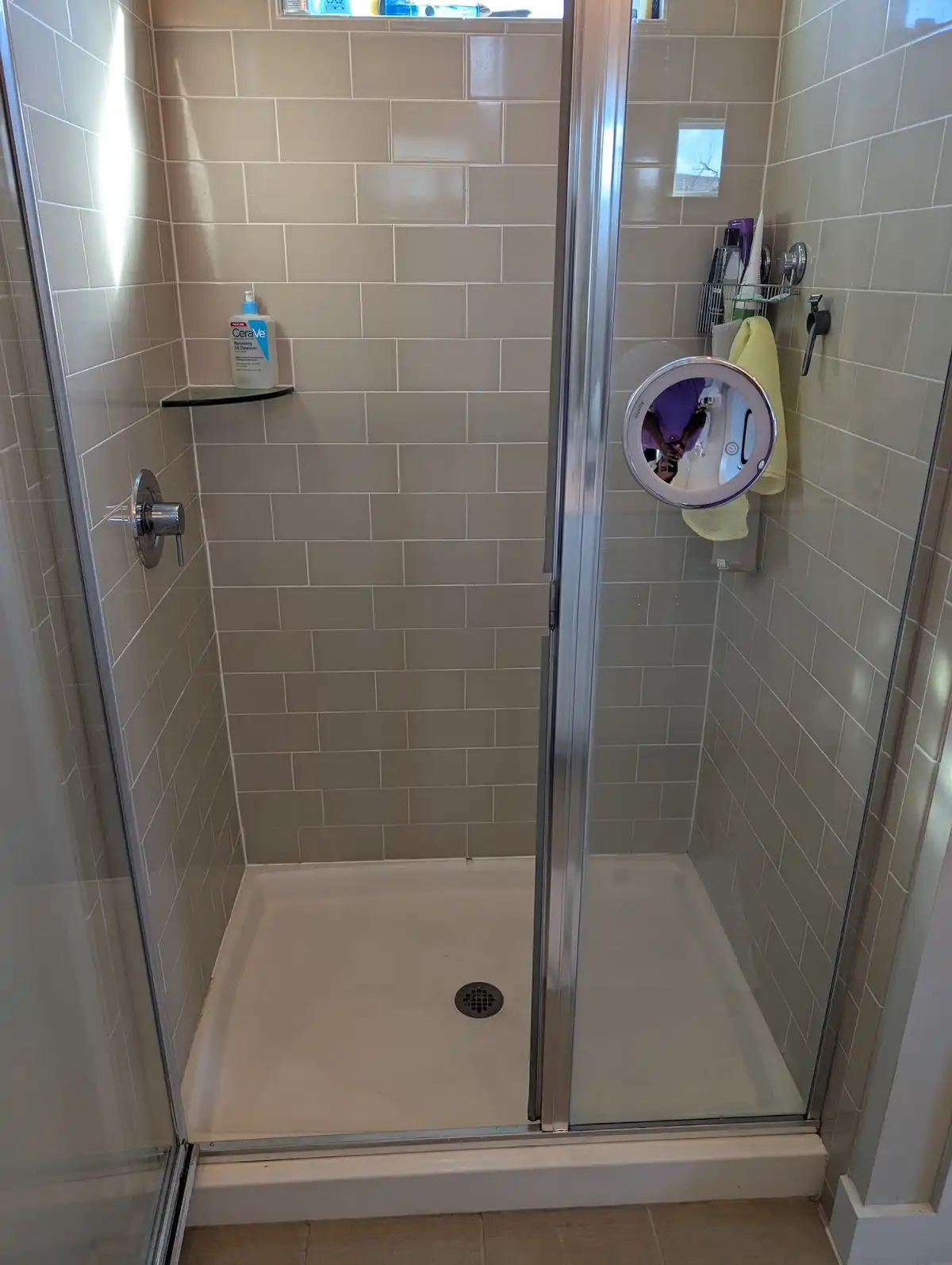 A photo of a clean, modern walk-in shower with beige subway tiles, a glass door, and a small shelf holding a CeraVe bottle…
