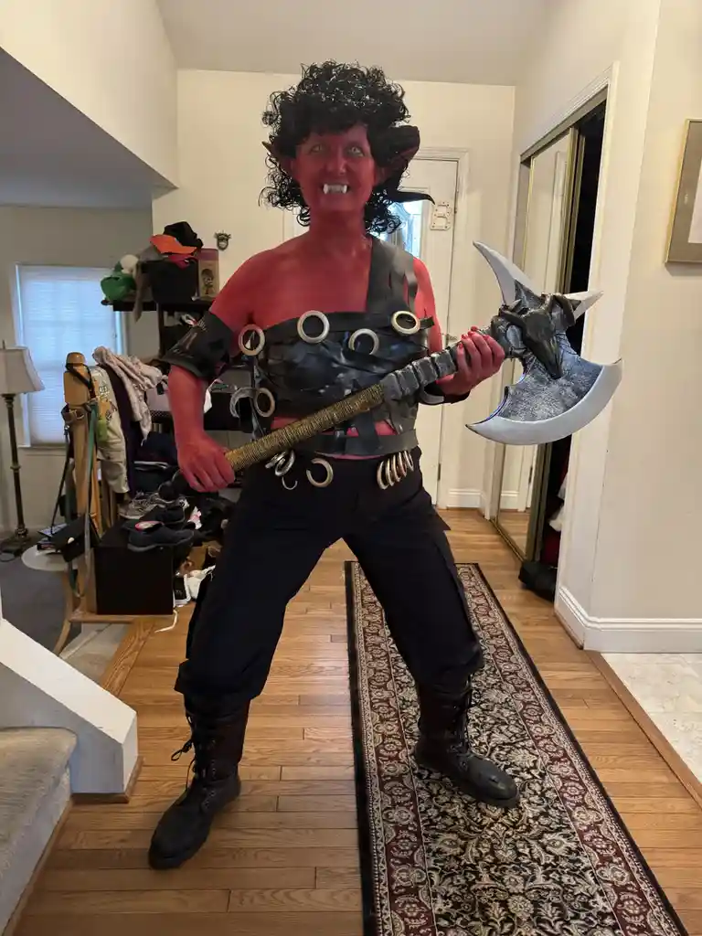 A person in a full demon costume with red face paint, black curly wig, pointed ears, fangs, and a black leather harness…