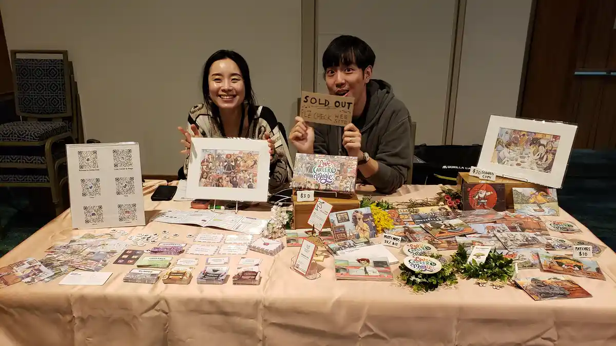 Two people sit behind a table covered in art prints, cards, and merchandise for 'Love, Career & Magic'. The man on the right…