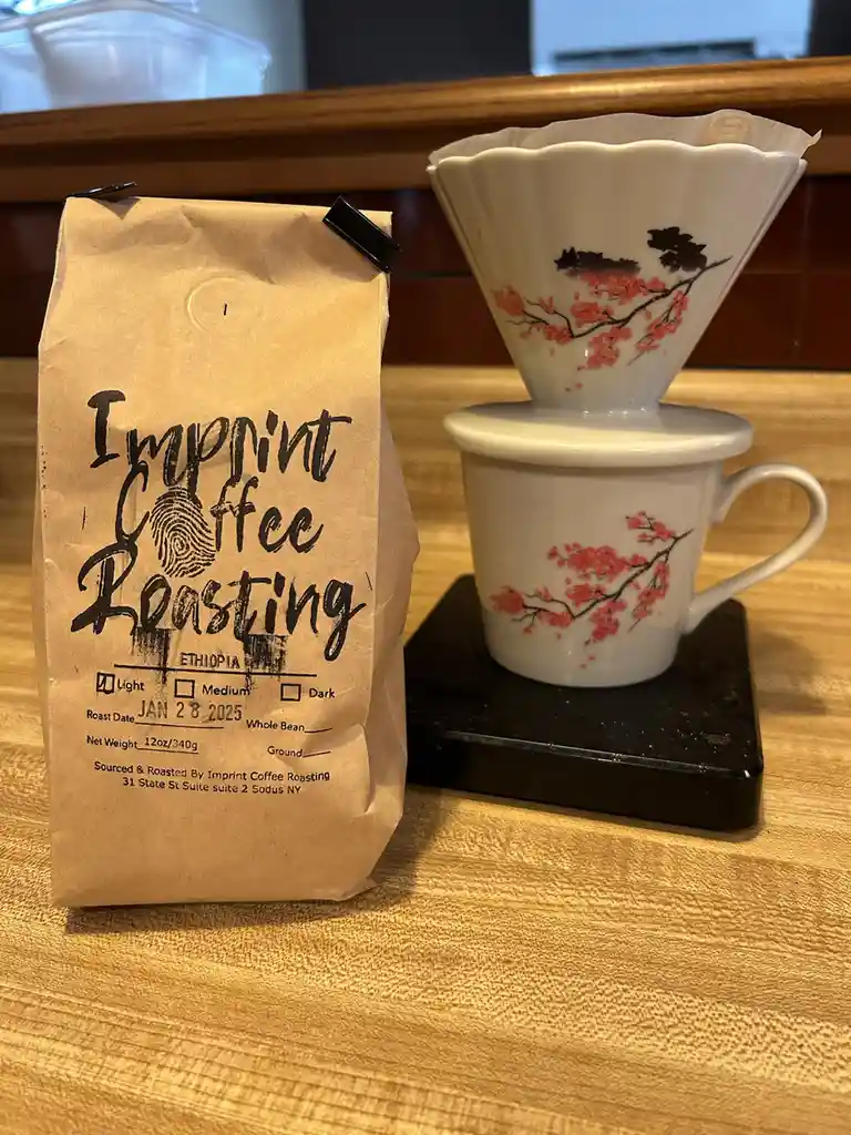 A brown paper bag labeled 'Imprint Coffee Roasting' sits next to a white ceramic pour-over coffee setup with cherry blossom…