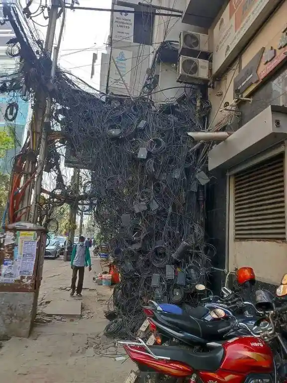 A chaotic tangle of black electrical wires, cables, and adapters hangs from a utility pole and covers a building facade in…