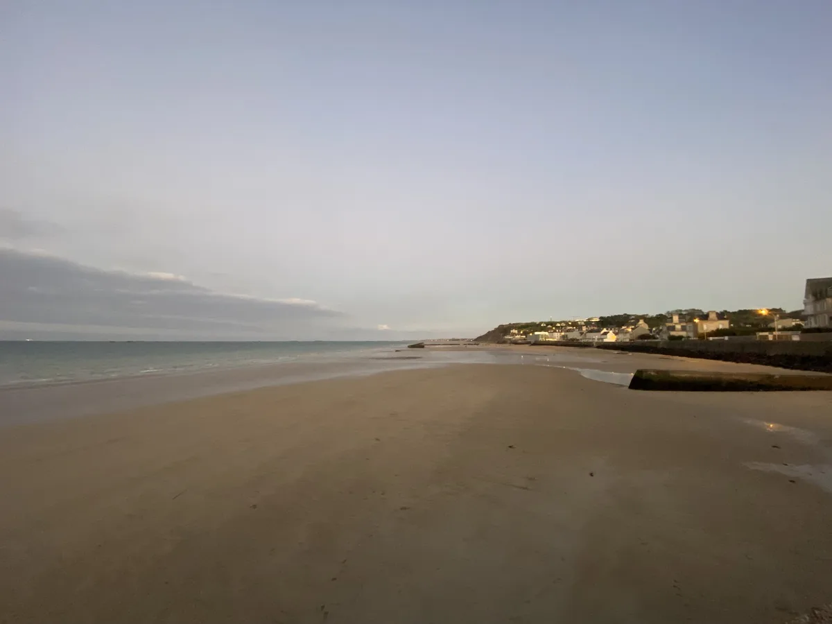 A wide, empty beach at dusk with calm water, wet sand, and a small coastal town with lit windows visible in the distance…