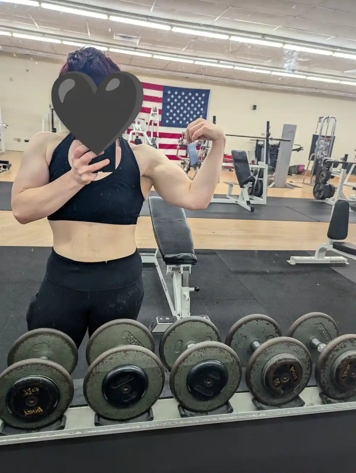 A person with a heart-shaped emoji covering their face takes a mirror selfie in a gym, flexing their bicep while wearing a…