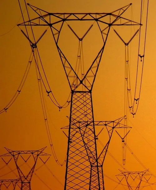 A silhouette of multiple electricity pylons against a warm orange sunset sky, with complex networks of wires and insulators…