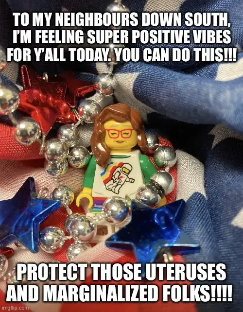 A close-up image of a Lego minifigure with brown hair and red glasses, wearing a white t-shirt with a rainbow and astronaut…