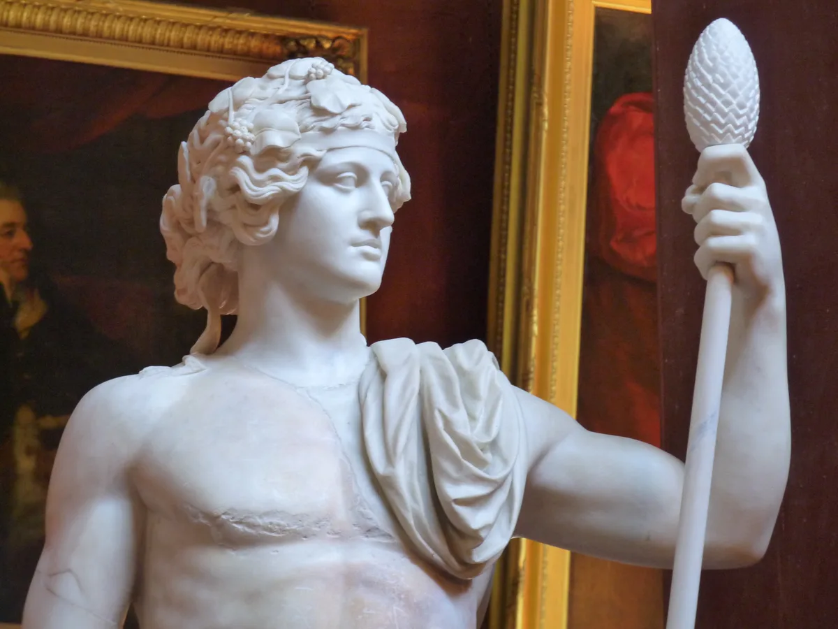 A marble statue of a classical male figure with curly hair adorned with grapevines and leaves, holding a staff topped with a…