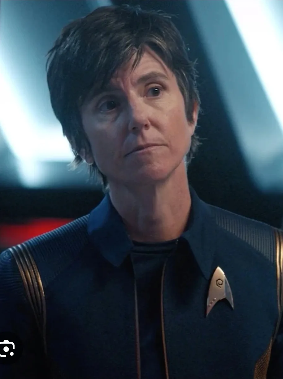 A woman with short dark hair, wearing a dark blue Starfleet uniform with gold trim and a captain's insignia, stares with a…